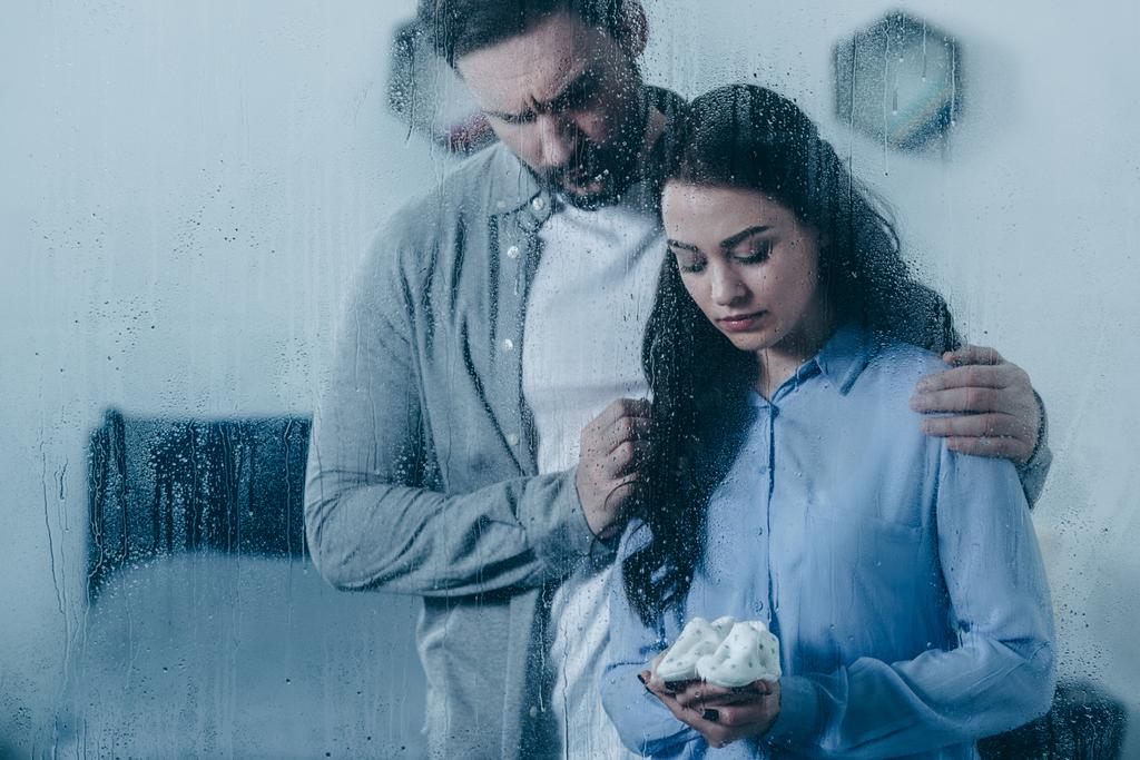 husband grieving and hugging wife with baby shoes through window with raindrops - Photo, Image