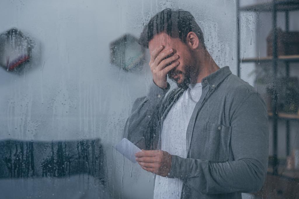 depressed man covering face with hand, holding photograph and crying through window with raindrops - Photo, Image