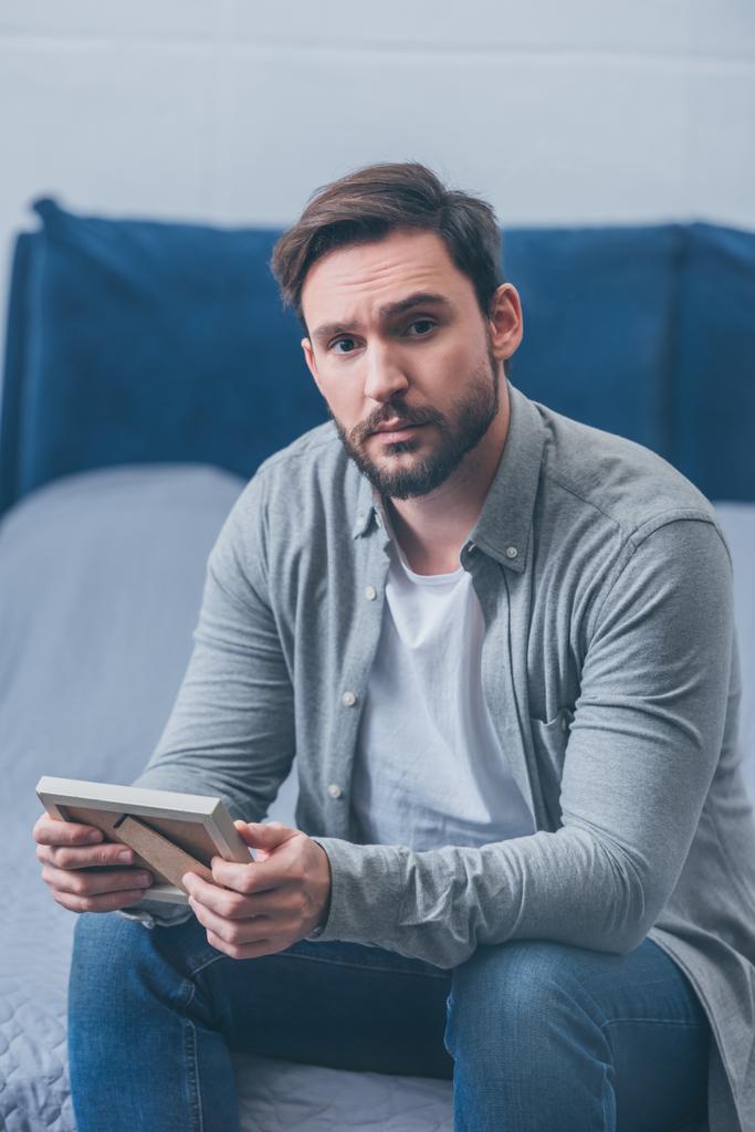 sad man sitting on bed, looking at camera and holding photo frame at home - Photo, Image