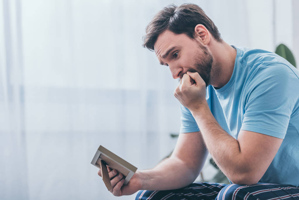 depressiver Mann sitzt, Mund mit der Hand bedeckt und Fotorahmen zu Hause betrachtet - Foto, Bild
