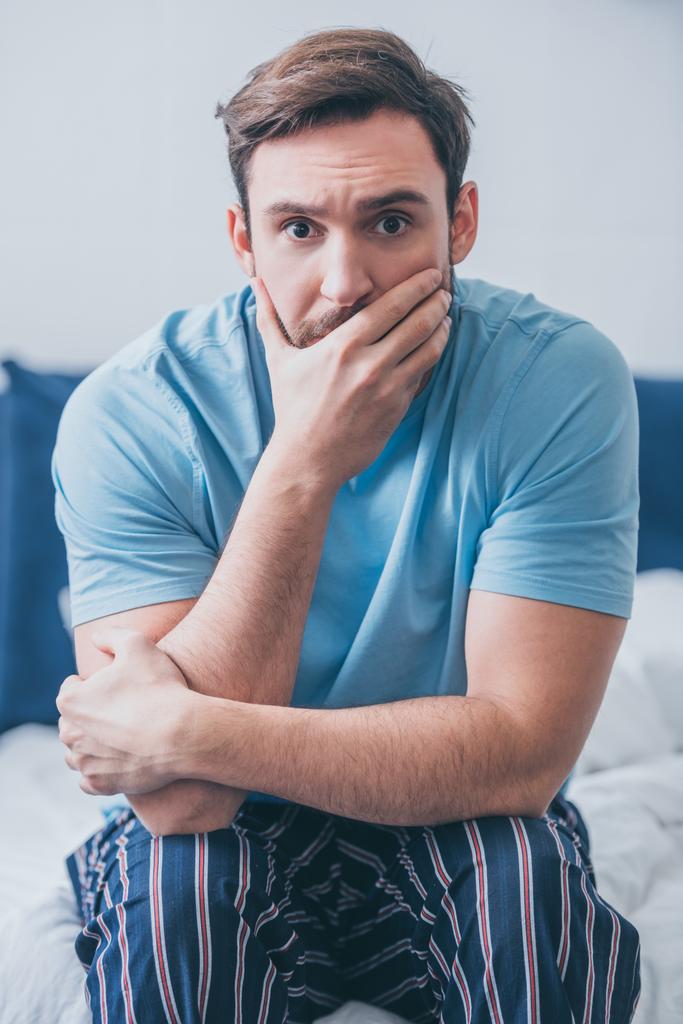 стресова людина сидить на ліжку, дивлячись на камеру і прикриваючи рот рукою вдома
 - Фото, зображення