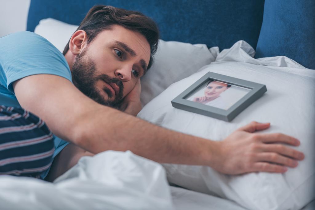 man in bed liggen en verdriet in de buurt van foto van vrouw op kussen thuis - Foto, afbeelding