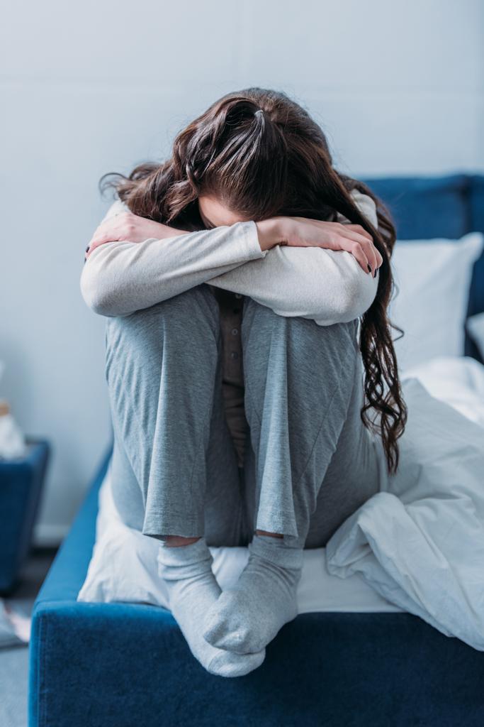 depressieve vrouw met hoofd naar beneden knuffelen knieën en zittend op bed thuis - Foto, afbeelding