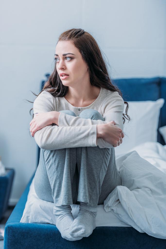 расстроенная женщина обнимает колени, сидит на кровати дома и смотрит в сторону
 - Фото, изображение