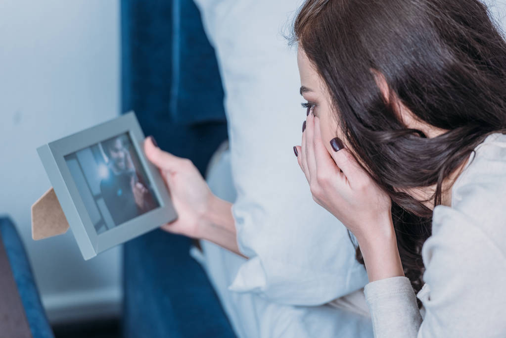 upset woman lying in bed, crying and looking at frame with picture of man at home - Photo, Image