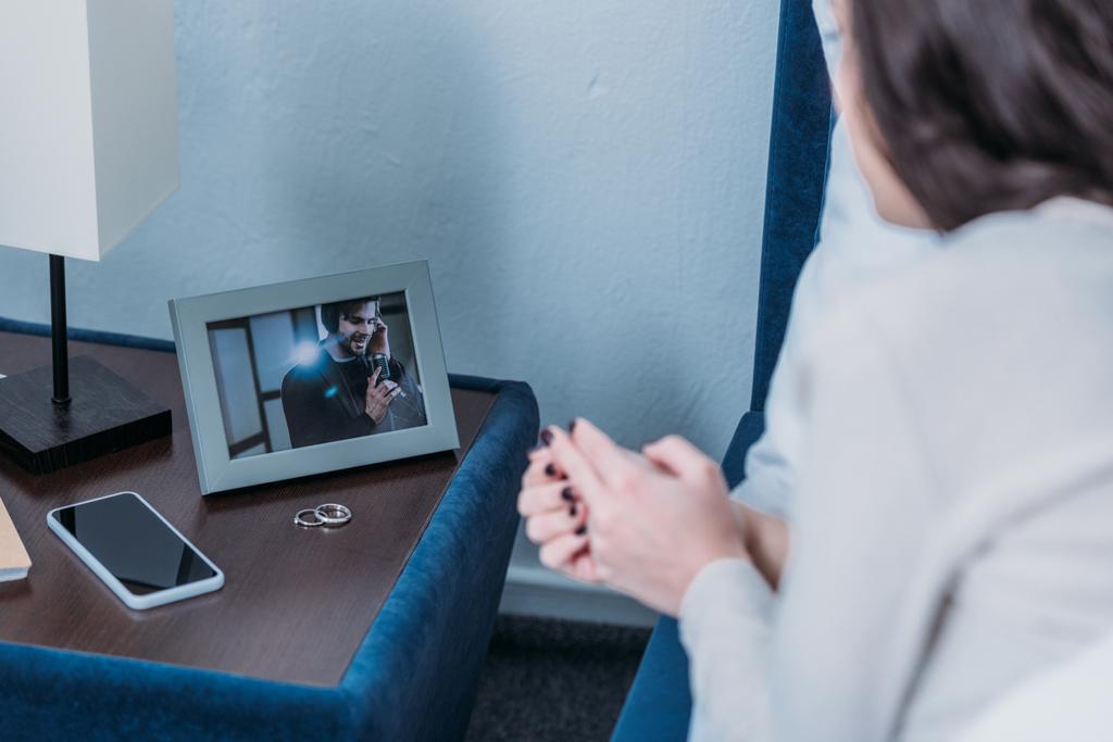 Ausgeschnittene Ansicht der Frau beim Betrachten des Rahmens mit Bild des Mannes und Trauringen auf Nachttisch  - Foto, Bild