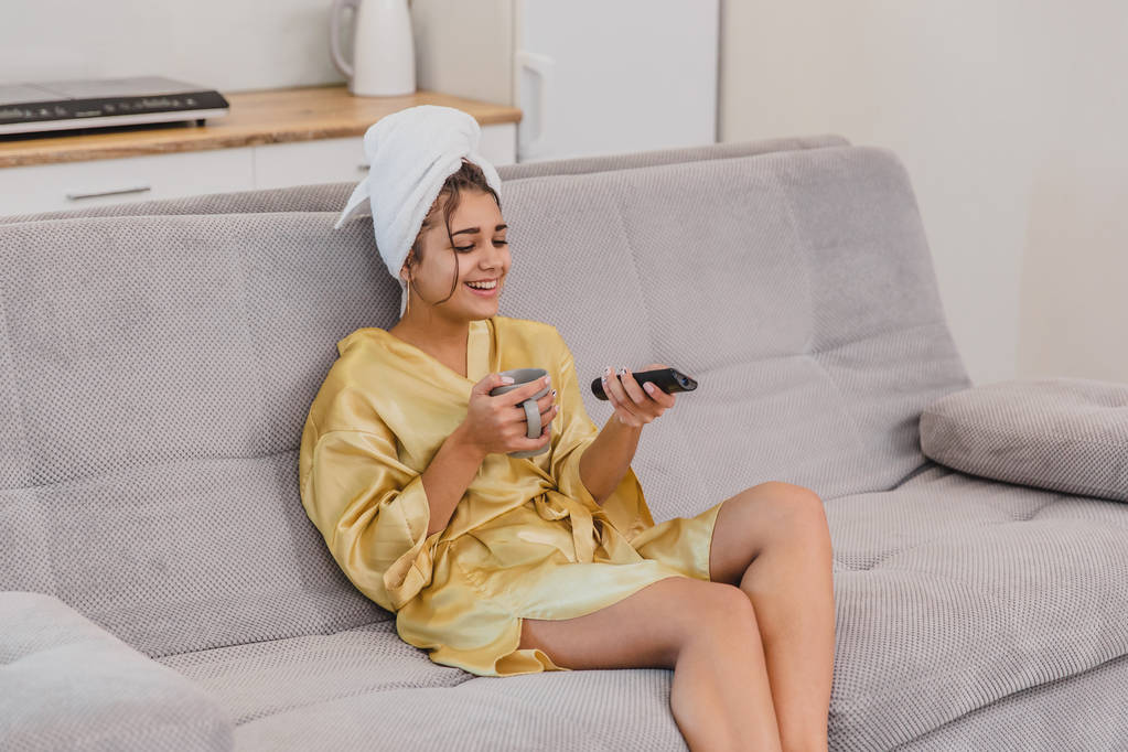Ευτυχισμένη γυναίκα με ένα τηλεχειριστήριο, επιλέγοντας ένα τηλεοπτικό κανάλι κατά τη διάρκεια μια ανάπαυση μετά το μπάνιο. Στο κεφάλι είναι μια πετσέτα. - Φωτογραφία, εικόνα