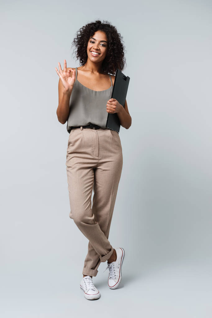 Full length of a happy young african woman casually dressed standing isolated over gray background, holding a notepad - Photo, Image