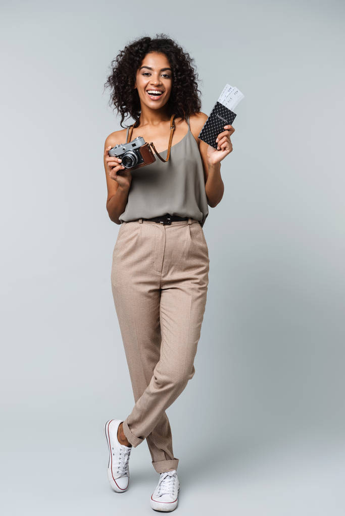 Longitud completa de una joven mujer africana feliz casualmente vestida de pie aislada sobre fondo gris, sosteniendo cámara fotográfica, mostrando pasaporte con boletos de avión
 - Foto, Imagen