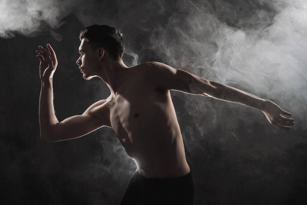 Un joven bailarín de ballet masculino con leggins negros y un torso desnudo realiza movimientos de baile sobre un fondo grunge gris, con una luz de luces y humo
. - Foto, imagen