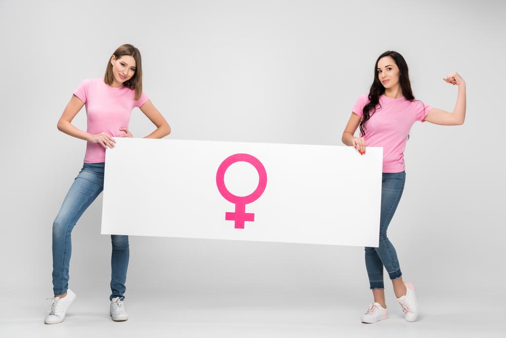 beautiful woman showing muscle and holding large sign with female symbol with friend on grey background - Photo, Image