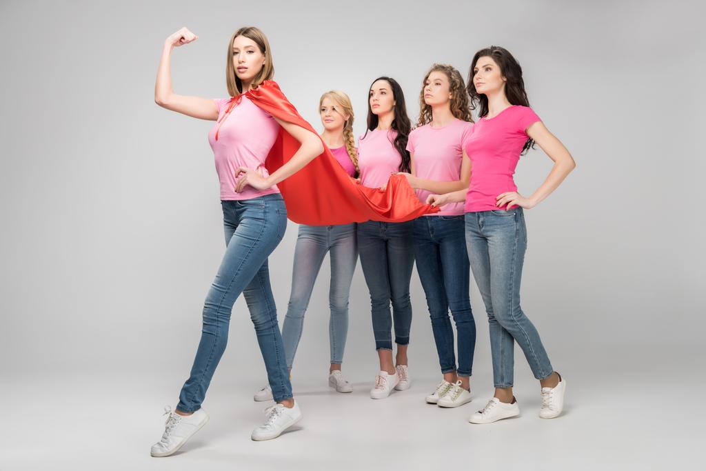 уверенная девушка в красном плаще супергероя показывает мышцы рядом с привлекательными женщинами на сером фоне
  - Фото, изображение
