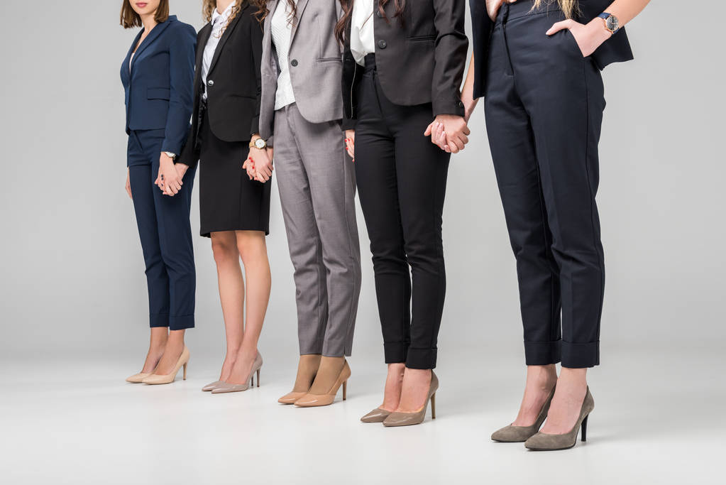обрізаний вид на жінок, що тримаються за руки і стоять в туфлях на високому каблуці на сірому фоні
 - Фото, зображення
