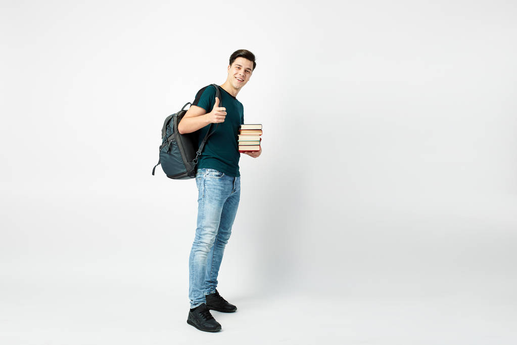 Забавный темноволосый парень с черным рюкзаком на плече, одетый в темную футболку и джинсы держит книги в руках на белом фоне
 - Фото, изображение