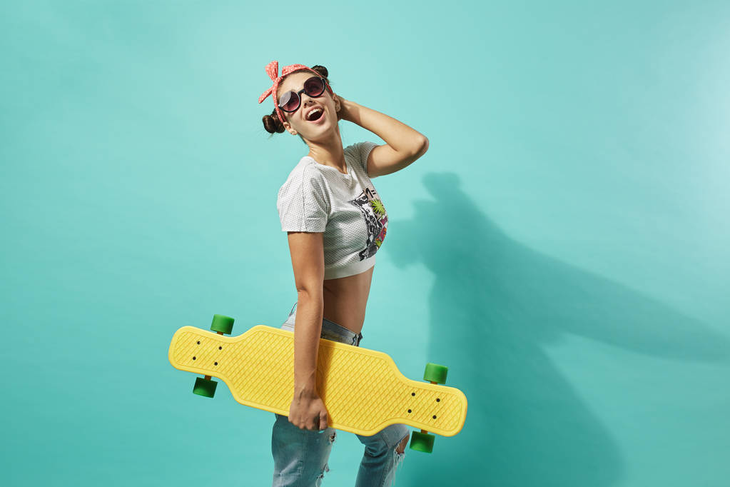 Engraçado jovem em óculos de sol e laço rosa na cabeça vestida de jeans e top stands com skate amarelo na parte de trás no fundo azul no estúdio
 . . - Foto, Imagem