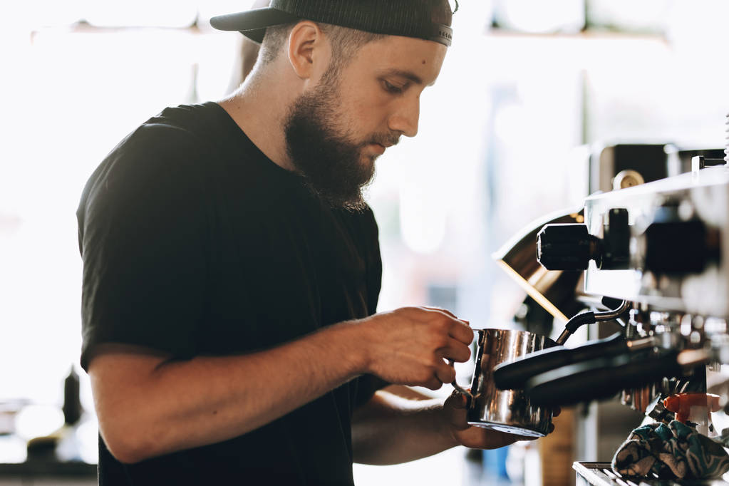 カジュアルな服を着て、髭、スタイリッシュな若い男料理モダンなコーヒー ショップでコーヒー マシンでコーヒー. - 写真・画像