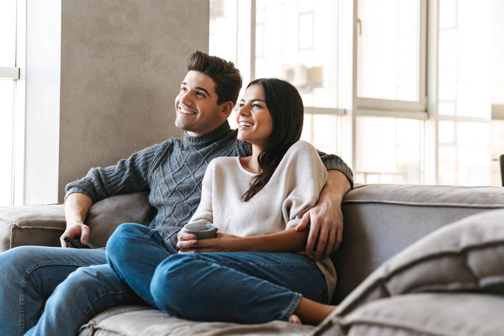 Happy νεαρό ζευγάρι κάθεται σε έναν καναπέ στο σπίτι, βλέποντας τηλεόραση και πίνοντας τσάι - Φωτογραφία, εικόνα