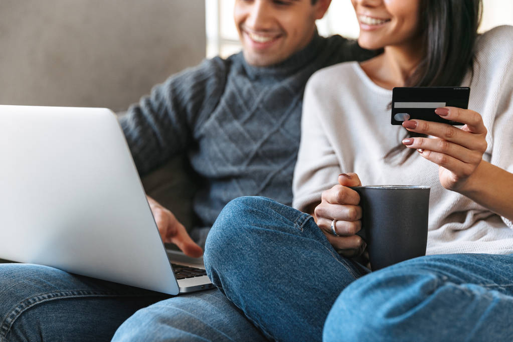 Щаслива молода пара сидить на дивані вдома, використовуючи ноутбук, показуючи кредитну картку
 - Фото, зображення
