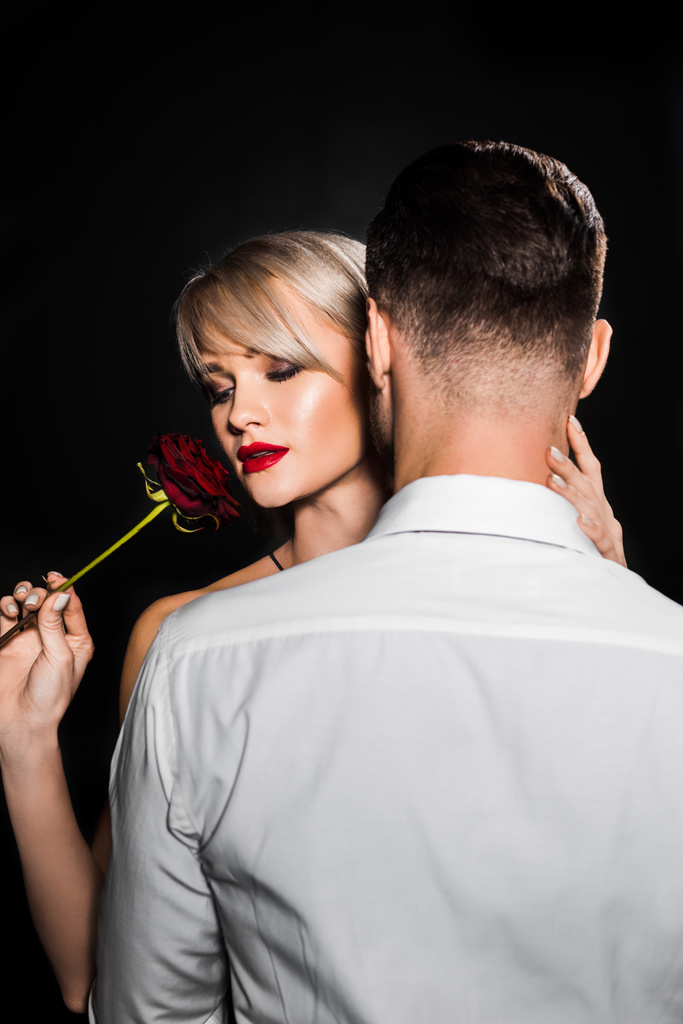 elegante blonde Frau hält rote Rose in der Hand und umarmt Mann, isoliert auf schwarz - Foto, Bild