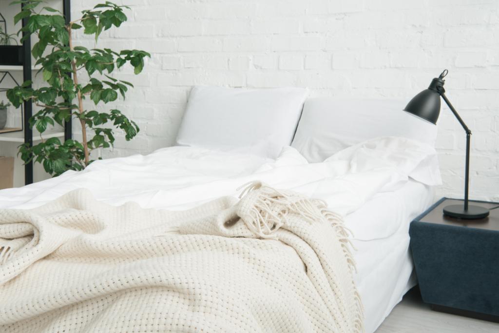 Bett mit weißer Decke und Kissen, Pflanze und Lampe auf schwarzem Nachttisch - Foto, Bild
