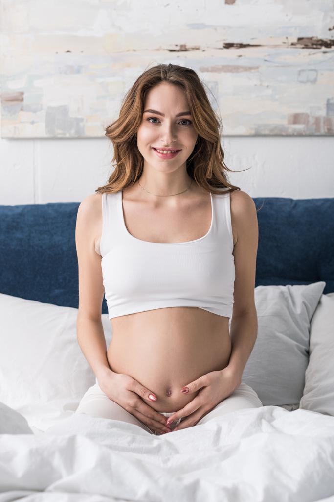陽気な妊娠中の女性のベッドの上に座っていると腹に触れる - 写真・画像