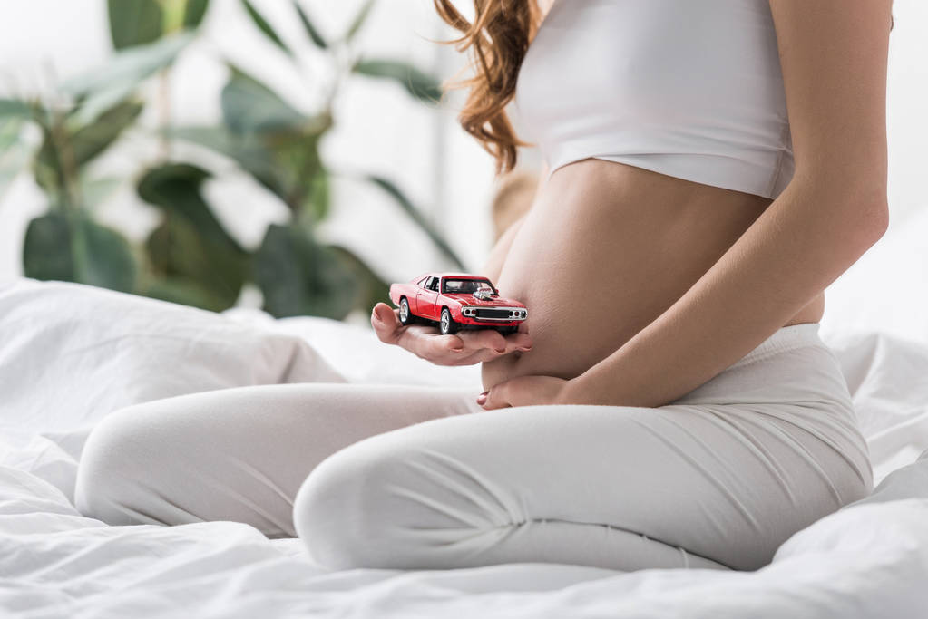 Vista parcial de la mujer embarazada sosteniendo coche de juguete rojo
 - Foto, Imagen