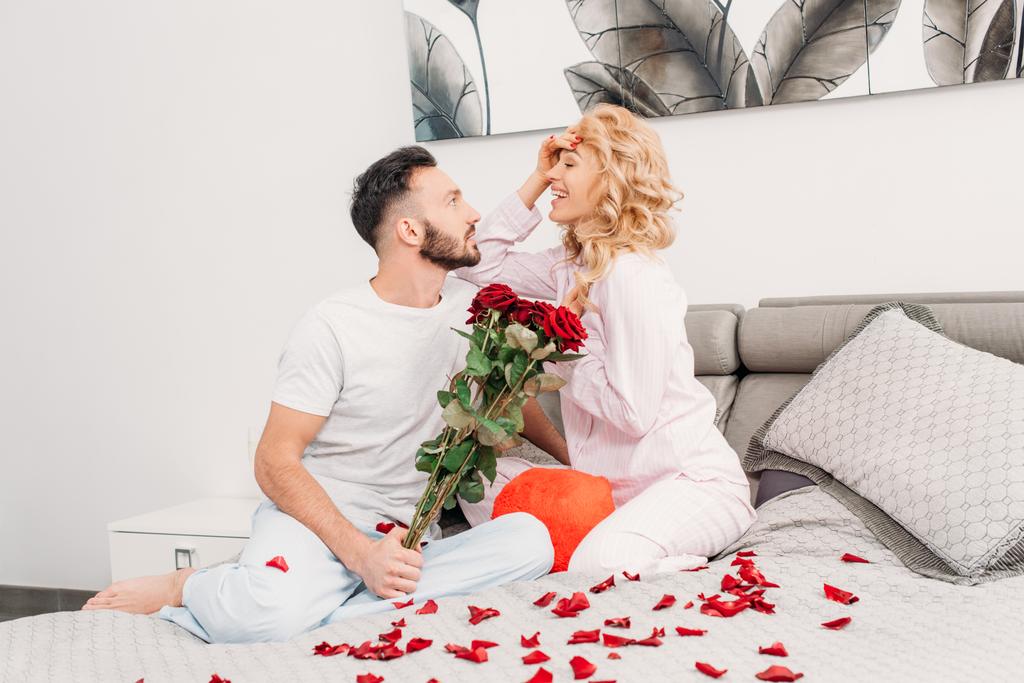Sonriente pareja romántica sentada en la cama con rosas rojas
 - Foto, imagen