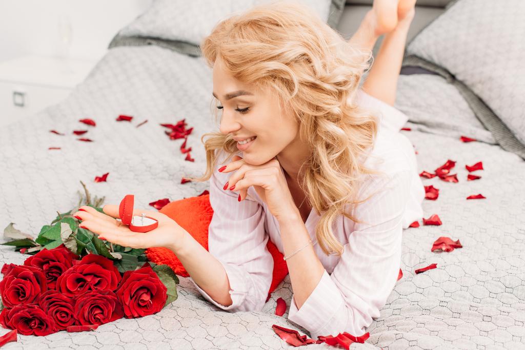 Szczęśliwy blond dziewczyna leżąc na łóżku z kwiatami i przytrzymując pierścień - Zdjęcie, obraz
