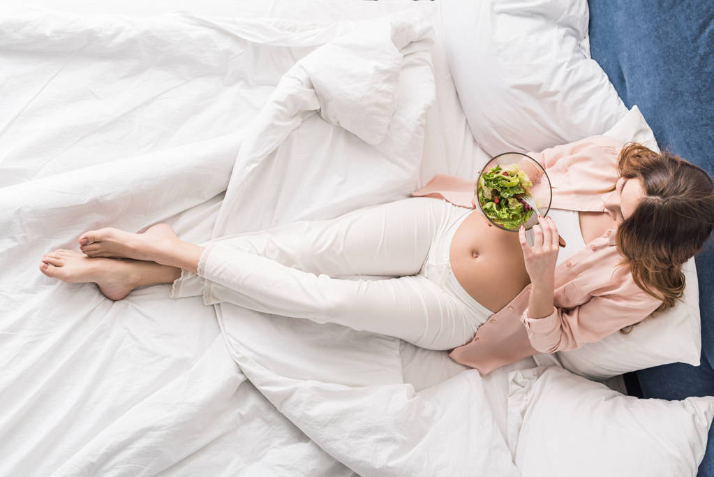 Top näkymä raskaana oleva nainen syö salaattia sängyssä
 - Valokuva, kuva