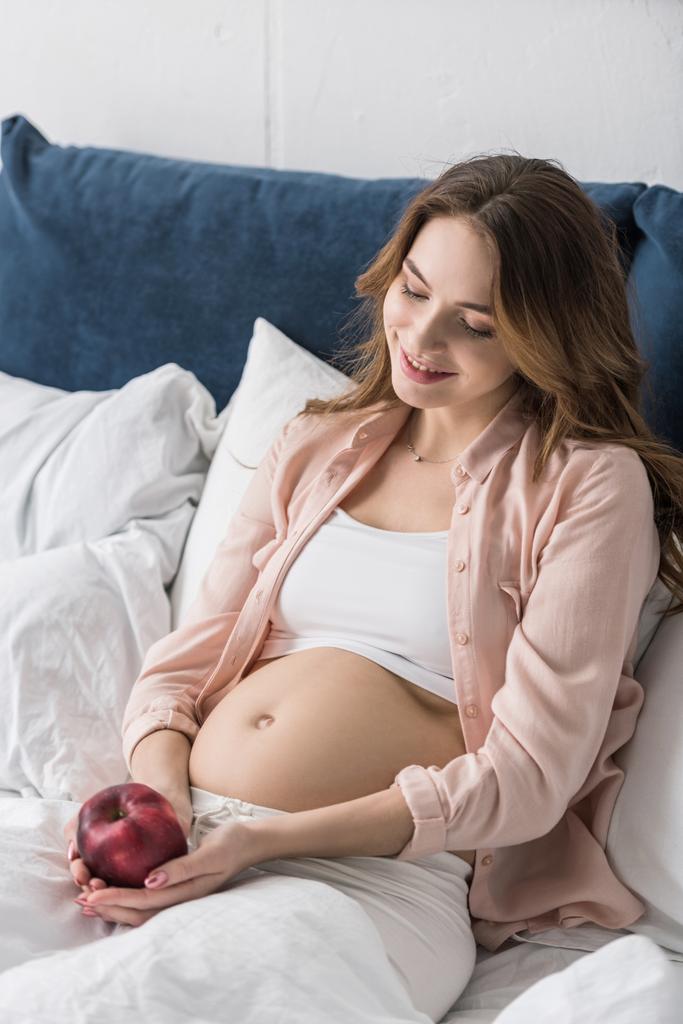 Femme enceinte souriante couchée au lit avec une pomme rouge
 - Photo, image