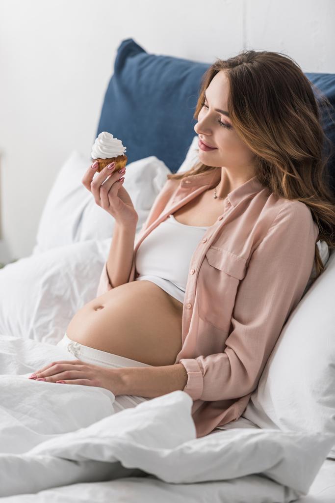 Hermosa mujer embarazada acostada en la cama y sosteniendo cupcake
 - Foto, imagen