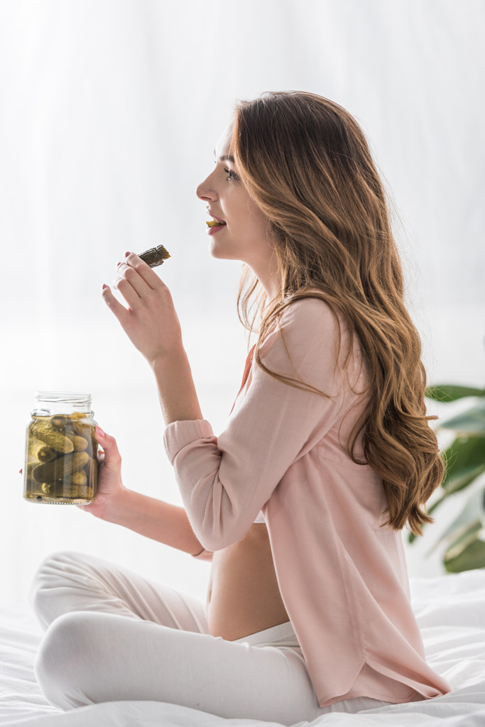Side view of long-haired pregnant woman eating pickles on bed - Photo, Image