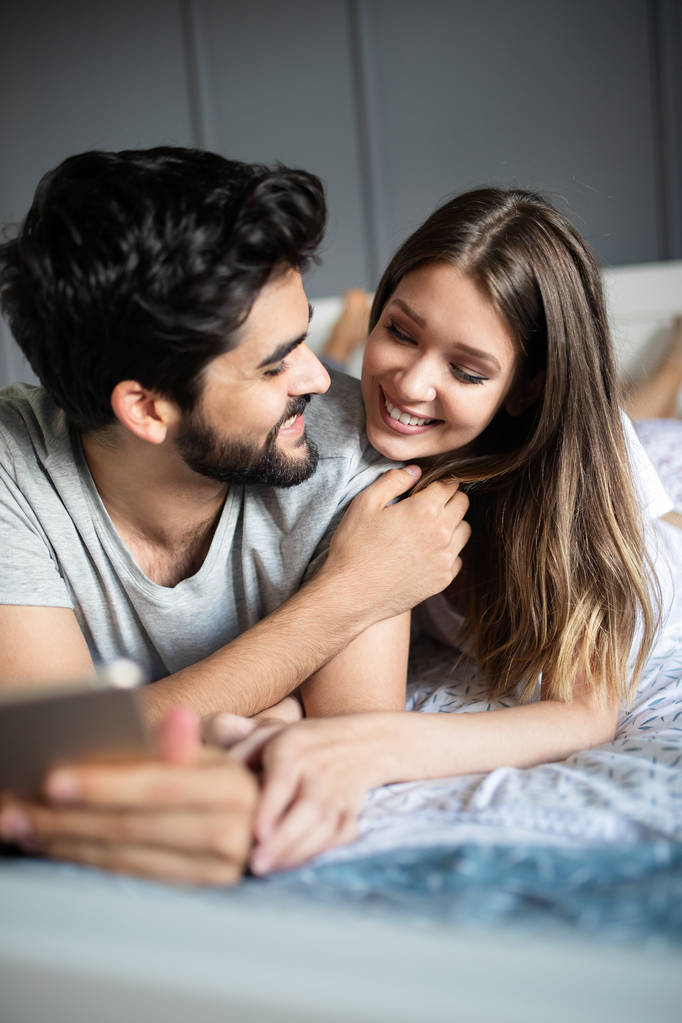 Fröhliches Paar schaut gemeinsam auf ein Tablet und lacht, während es auf dem Bett liegt - Foto, Bild