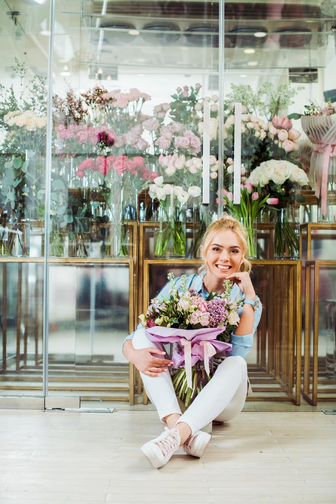 beautiful smiling woman holding flower bouquet, looking at camera and sitting in front of flower shop  - Photo, Image