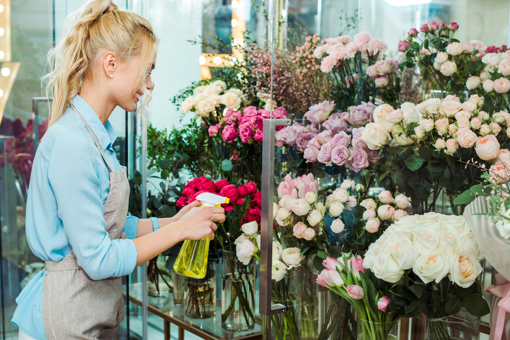 vrouwelijke bloemist in schort bloemen spuiten met sproeiflacon in bloemenwinkel - Foto, afbeelding