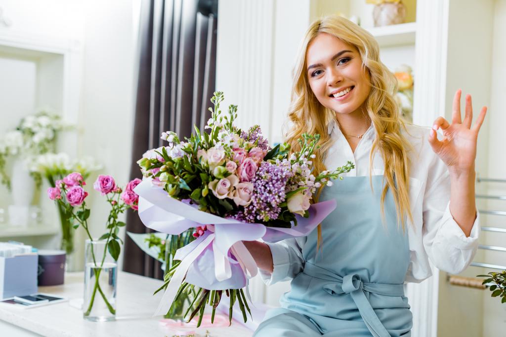 belle femme souriante propriétaire de magasin de fleurs assis sur le comptoir et tenant bouquet tout en montrant ok signe
 - Photo, image