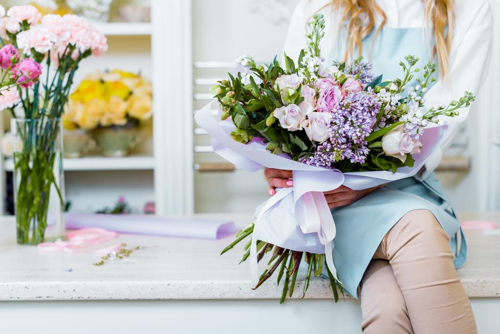cropped view of female florist sitting on counter and holding bouquet with lilac and roses in flower shop - Photo, Image