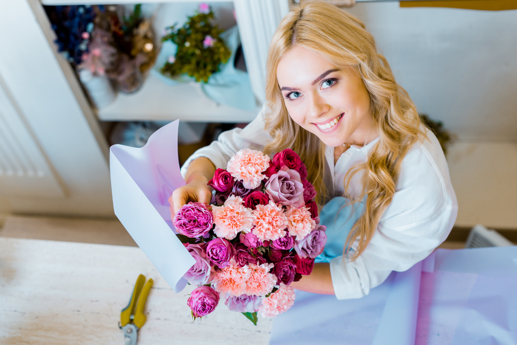 schöne Floristin blickt in die Kamera, während sie einen Strauß mit Rosen und Nelken im Blumenladen hält  - Foto, Bild