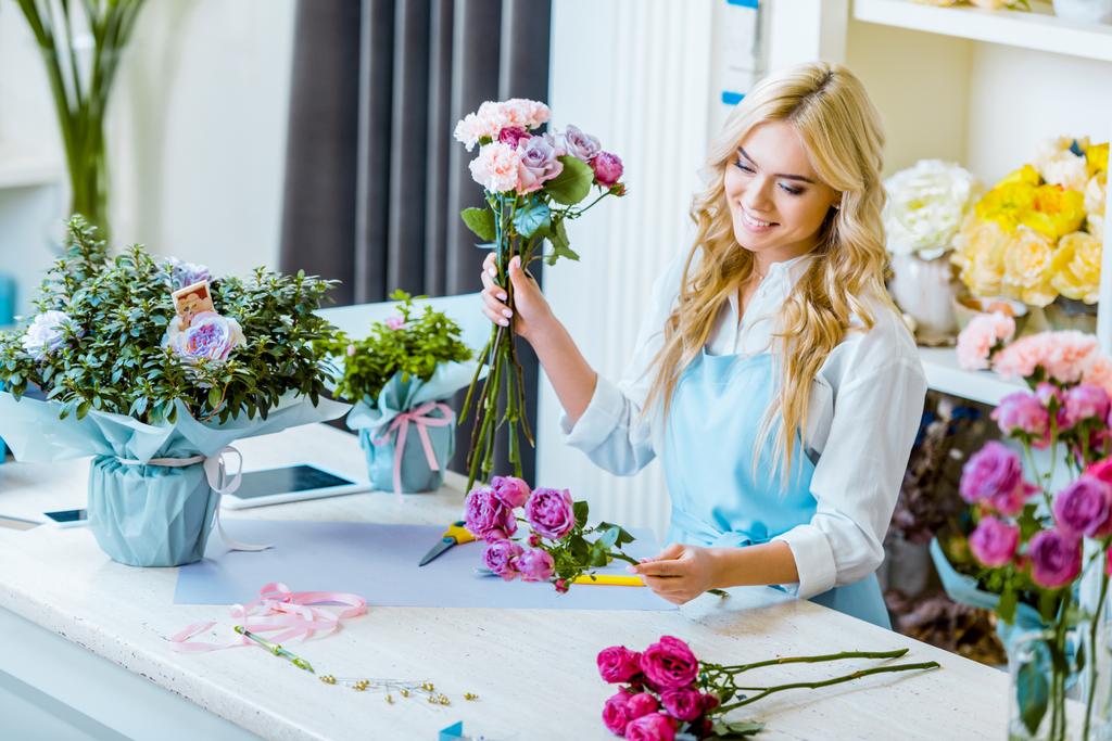 красивий усміхнений жіночий флорист в фартусі розставляє букет з трояндами в квітковому магазині
  - Фото, зображення