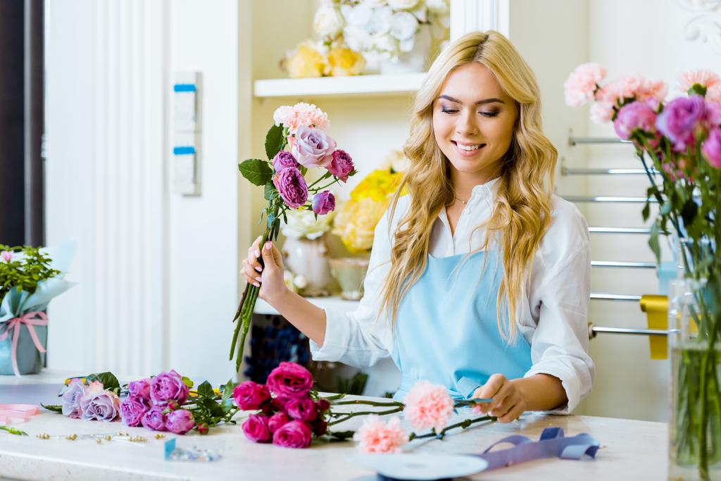 привлекательная флористка в фартуке, раскладывающая букет с розами в цветочном магазине
  - Фото, изображение
