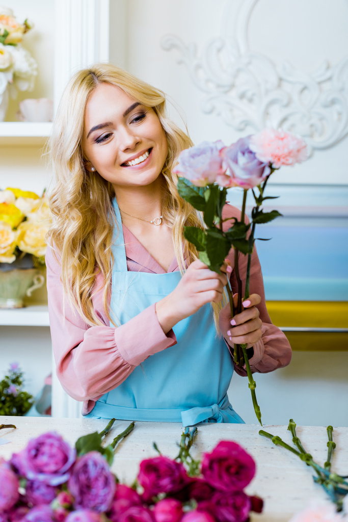 красива щаслива жінка-флорист в фартуху, що влаштовує букет з трояндами в квітковому магазині
  - Фото, зображення