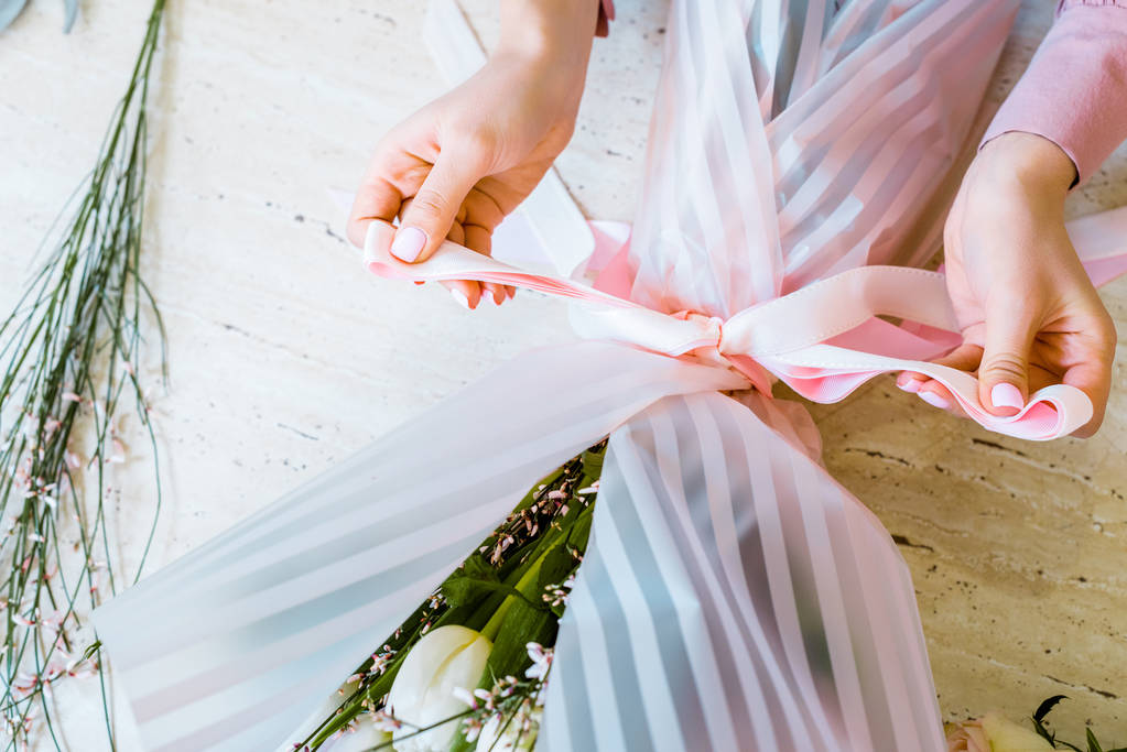 vista parcial de la floristería femenina atando arco con cinta mientras envuelve ramo de flores
  - Foto, Imagen