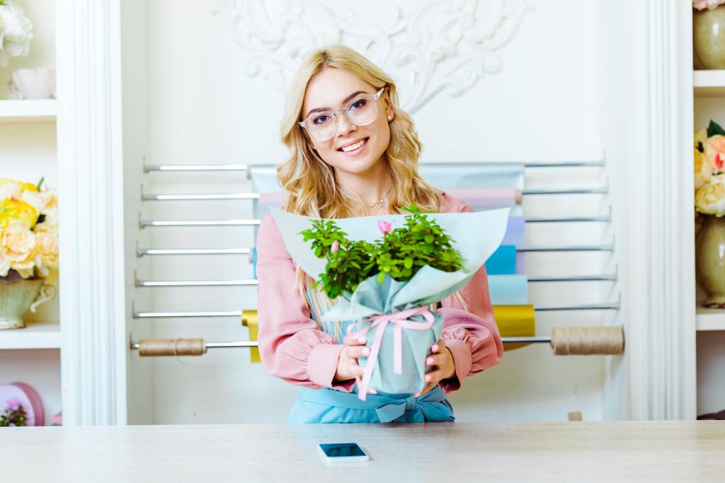 bela florista feminina em óculos olhando para a câmera e apresentando buquê na loja de flores
 - Foto, Imagem
