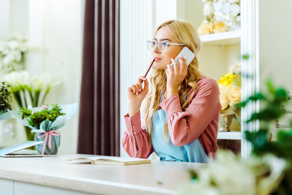hermosa mujer propietaria de la floristería en gafas hablando en el teléfono inteligente y sosteniendo lápiz
 - Foto, imagen
