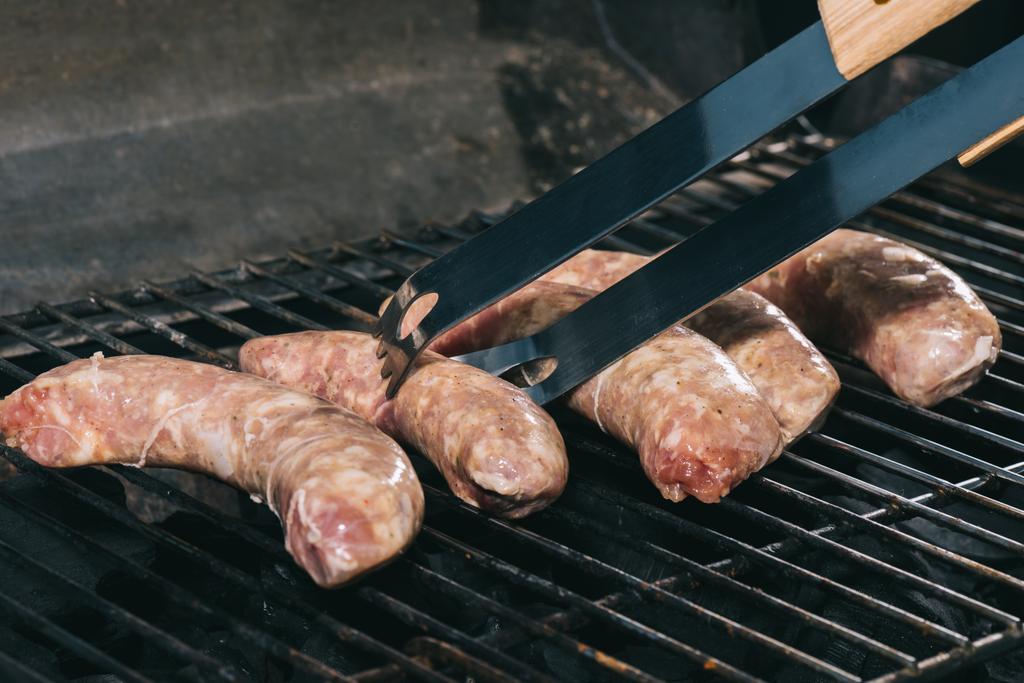 gros plan des pinces à épiler près des saucisses fraîches non cuites sur les grilles de barbecue
 - Photo, image