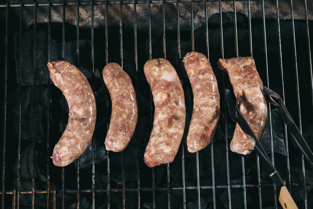 bovenaanzicht pincet in de buurt van ongekookte verse worst op BBQ-grill roosters - Foto, afbeelding