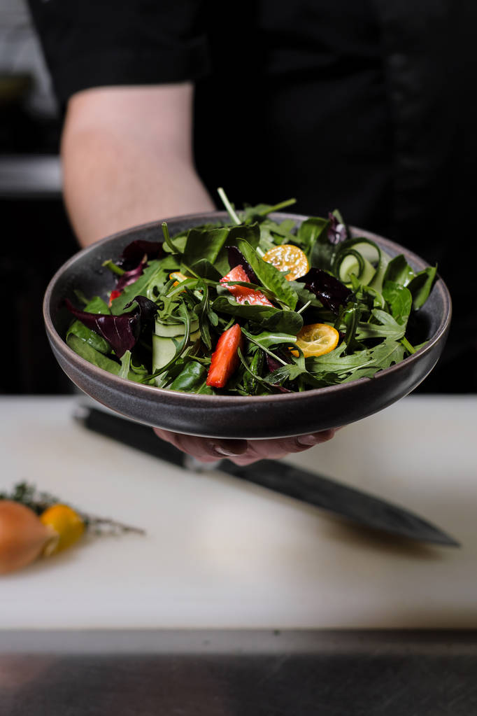 Szakács férfi fekete kötény egy tál saláta tart a kezében. A háttérben az a konyha, az étterem. - Fotó, kép