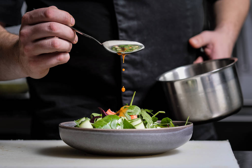 Close-up das mãos de um chef masculino sobre um fundo preto. Despeje o molho da colher no prato de salada
. - Foto, Imagem