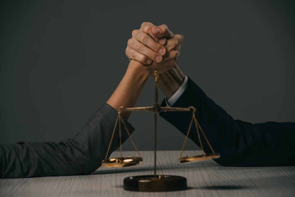 partial view of businessman and businesswoman arm wretsling on wooden table with scales of justice on grey - Photo, Image