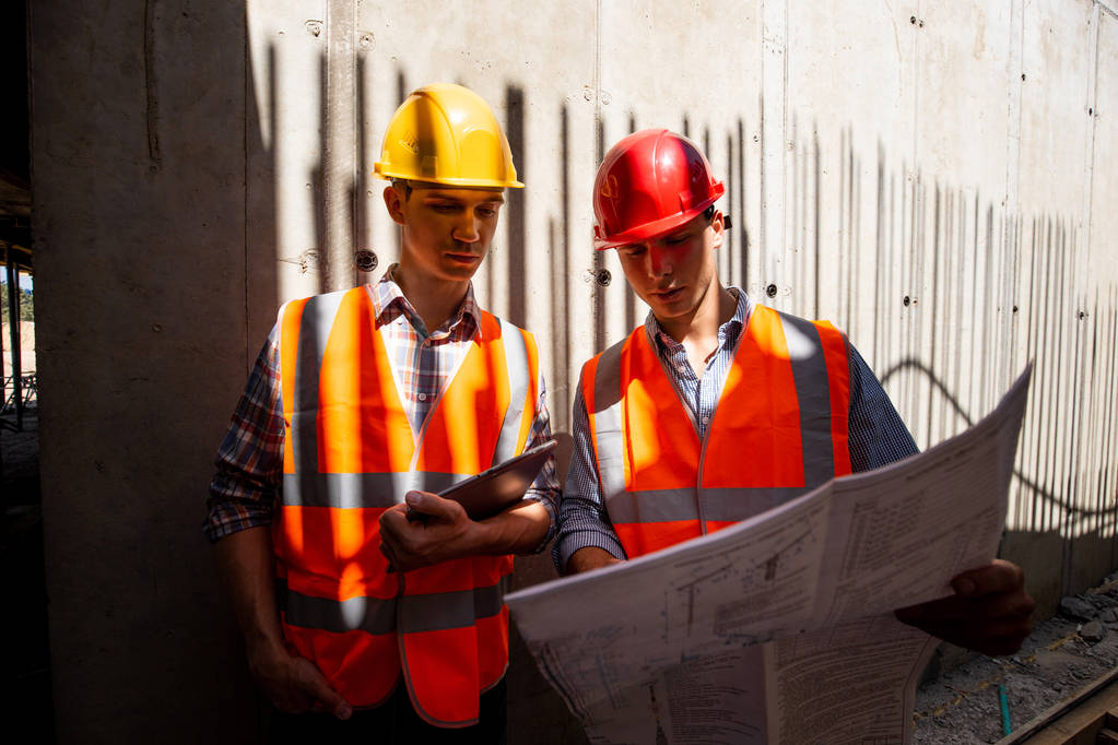 Dwóch inżynierów budownictwa ubrany w pracy pomarańczowe kamizelki i kaski Przejrzyj dokumentację projektu na placu budowy - Zdjęcie, obraz